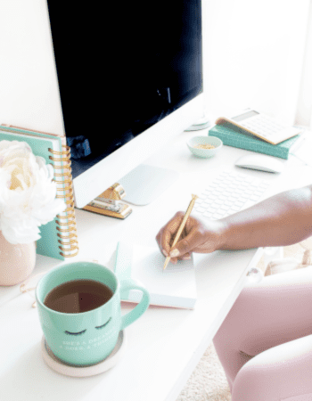 woman writing in notepad in front of computer with cup of coffee next to her and other books on the desk